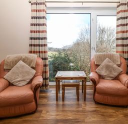A sitting area with two leather armchairs, cushions, a wooden table, striped curtains, and a large window with a view of trees at Bron Aber in Arthog near Dolgellau