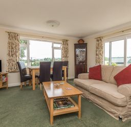 A living room with a beige sofa with red cushions a wooden coffee table a wooden dining table with four black chairs a television on a wooden stand and large windows with floral curtains at Pipistrelle Lodge in Plymtree