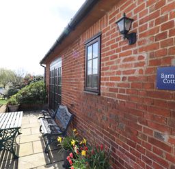 An outdoor patio area with a bench and table next to a brick wall with a sign that reads Barn Owl Cottage with flowers and a lawn by water at Barn Owl Cottage in Little Glemham near Wickham Market