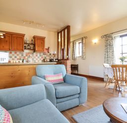 A living area with blue armchairs a wooden kitchen with cabinets a dining table with chairs and large windows at Barn Owl Cottage in Little Glemham near Wickham Market