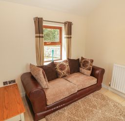 A small sitting area with a sofa cushions window with curtains a wooden side table and a radiator at Y Beudy Cottage in Clynnog Fawr
