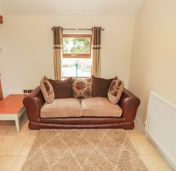 A living room with a brown leather and fabric sofa with cushions a small wooden side table a window with curtains and a radiator at Y Beudy Cottage in Clynnog Fawr