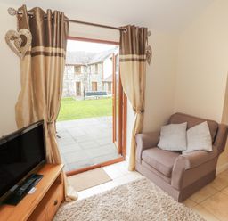 A small living room with a TV on a wooden stand an armchair with two cushions and glass doors with curtains leading to a garden at Y Beudy Cottage in Clynnog Fawr