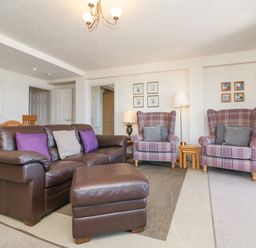 A living room with a brown leather sofa and ottoman two plaid armchairs wooden dining chairs and framed pictures on the walls at 37 Ocean Park in Westward Ho