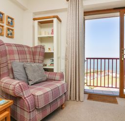 A chair with cushions next to a wooden side table and a white shelving unit by a sliding door with a view of houses and the sea at 37 Ocean Park in Westward Ho