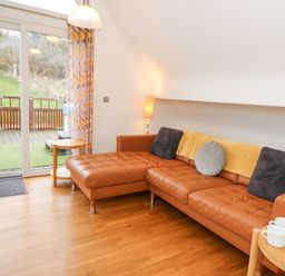 A living room with a brown leather sectional sofa with cushions and a throw next to a sliding door leading to a deck with outdoor furniture at Ty Henri in Tresaith
