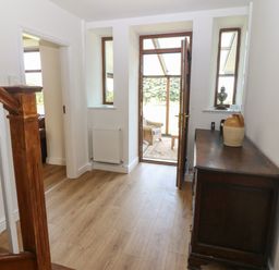 A hallway with wooden stairs on the left a dark wooden cabinet on the right and an open door leading to a sunroom at Y Berth Ddu Farmhouse in Clynnog Fawr