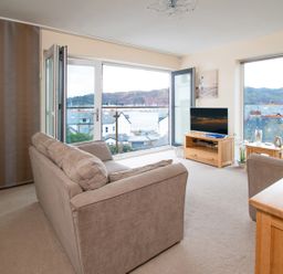 A living room with beige sofas a wooden TV stand and large windows with a view of houses and hills at The Moorings in Deganwy near Llandudno
