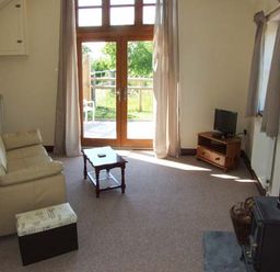 A living room with a beige sofa small wooden coffee table television on a wooden stand wood burning stove and glass doors at Dovetail in Westbury-on-Severn