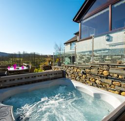 An outdoor hot tub next to a stone wall and glass railing with a patio dining set and barbecue grill in the background at Browside Little Arrow near Coniston