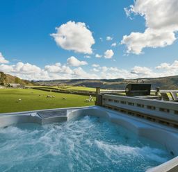 An outdoor hot tub with bubbling water overlooking a green field with sheep and hills in the background at Browside Little Arrow near Coniston