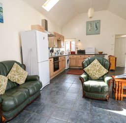 A living area with green leather sofas and a wooden table next to a kitchen with wooden cabinets and white appliances at Yr Hen Ysgubor Trelech near St Clears