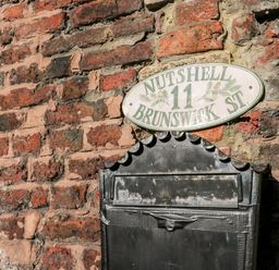 A black metal mailbox attached to a brick wall with an oval white sign above it saying Nutshell 11 Brunswick St