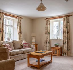 A sitting room with a beige sofa two windows with floral curtains wooden coffee and side tables with lamps and a floral basket at Crabtree Hawkshead