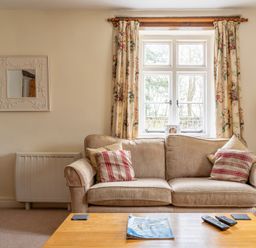 A beige sofa with patterned cushions in front of a window with floral curtains and a wooden coffee table with remotes and a magazine in Crabtree Hawkshead