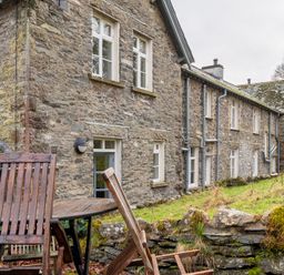 A stone house with multiple windows and a grassy yard with wooden chairs and a table at Forge in Graythwaite near Hawkshead