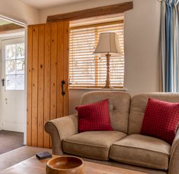 A living room with a beige sofa with red cushions a wooden coffee table a lamp by the window and a wooden door at Newlands in Graythwaite near Hawkshead