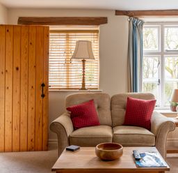 A living room with a beige sofa with red cushions a wooden coffee table with a bowl and a remote control a wooden side table with a lamp and plant wooden blinds and windows with blue curtains at Newlands in Graythwaite near Hawkshead