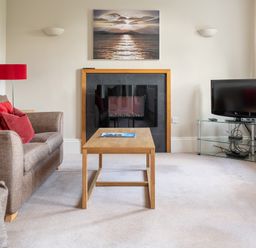 A living room with a sofa red pillows a wooden coffee table fireplace and a television on a glass stand at Mill Cottage in Graythwaite near Hawkshead