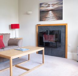 A living room with a sofa red cushions a wooden coffee table a wall-mounted fireplace a red lamp and a TV on a glass stand at Mill Cottage in Graythwaite near Hawkshead