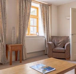 A room with two windows with curtains a wooden side table with a vase a brown armchair an open door and a wooden coffee table with a magazine Mill Cottage Graythwaite near Hawkshead
