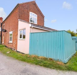 A brick house with a blue wooden fence and a small patch of grass near a road at 2 Lock Cottages in Honing near East Ruston