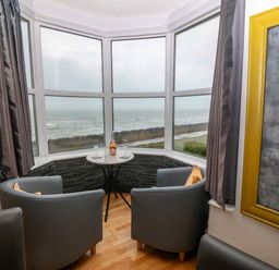 A corner seating area with two grey chairs and a small round table with glasses and a bottle by a large window overlooking the sea at Morolwg in Llanaber near Barmouth