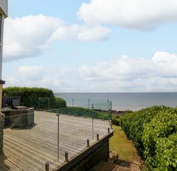 A wooden deck with glass railing overlooking the sea and a green hedge at Morolwg in Llanaber near Barmouth