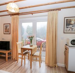 A dining area with a small wooden table and two chairs beside glass doors with curtains a TV on a wooden stand and a microwave on a countertop at Woods Edge in Mitcheldean