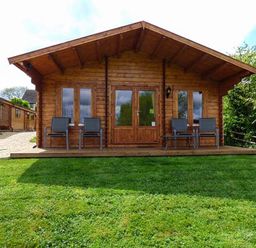 A wooden cabin with four chairs on the porch and a green lawn in front at Pennylands Willow Lodge in Broadway
