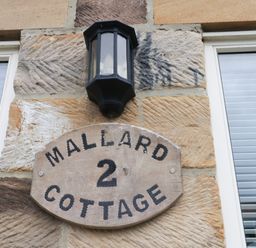 A wooden sign with the text Mallard 2 Cottage mounted on a stone wall under a black outdoor light fixture at Mallard Cottage in Whitby