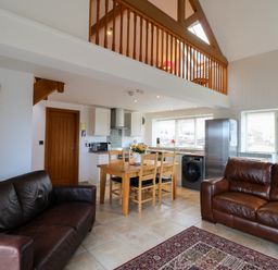An open plan living area with two brown leather sofas a wooden dining table with chairs kitchen appliances and a balcony railing above at Mallard Cottage in Whitby