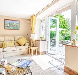 A living room with a sofa yellow cushions a small wooden table with a lamp an open door to a garden and a coffee table with pastries and mugs at Bwythyn Y Wennol in Nantmor near Beddgelert