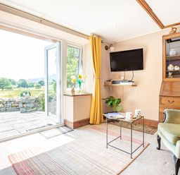 A living room with an open glass door leading outside a wooden cabinet a green armchair and a wall-mounted television at Bwythyn Y Wennol in Nantmor near Beddgelert 