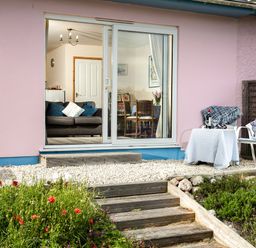 A patio area with a table and chairs outside a pink building with sliding glass doors showing a living room and dining area at Nectan in Mevagissey
