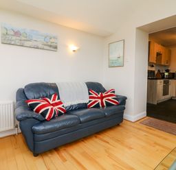A living room with a blue leather sofa with union jack pillows and a blanket next to a kitchen at The Well House Apartment in Watchet
