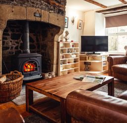 A living room with a stone fireplace and wood burning stove a wooden coffee table leather armchairs a TV and shelves at Bastle House in Haltwhistle near Hadrians Wall