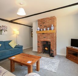 A living room with a wooden coffee table a blue sofa a brick fireplace with a wood stove and a TV on a wooden stand at Hadleigh Farm Cottage in Gayton