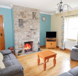 A living room with a stone fireplace a wooden coffee table grey sofas a small wooden TV stand with a television and patterned curtains at Tieve Baun in Ballintrillick near Kinlough County Sligo