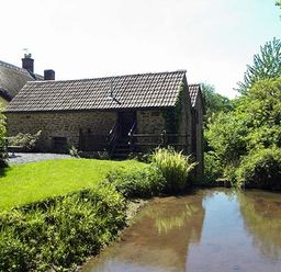 A stone building with a tiled roof near a pond with green grass and trees surrounding it at The Mill in Bicknoller