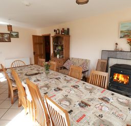 A dining room with a long table covered with a coffee-themed tablecloth surrounded by wooden chairs a wood-burning stove with a fire a wooden cabinet with flowers and framed pictures on the walls at Lough Mask Road Fishing Lodge in Ballinrobe County Mayo