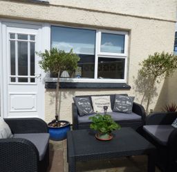 An outdoor seating area with black wicker furniture cushions pillows and potted plants against a beige wall at Spindrift in Porth near Newquay