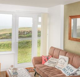 A living room with a brown couch with cushions a wooden coffee table a lamp and large windows with a view of grass and road at Spindrift in Porth near Newquay