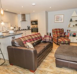 An open-plan kitchen and living room with a brown leather sofa, plaid armchair, ottoman, kitchen island with stools, and a boat-shaped bookshelf at The Old Laundry in Clappersgate near Ambleside