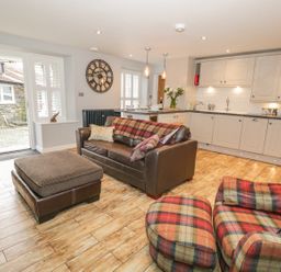 A kitchen and living area with a brown leather sofa, plaid armchair, ottoman, clock on the wall, and white cabinets at The Old Laundry in Clappersgate near Ambleside