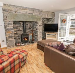 A living room with a stone fireplace a wood stove a plaid armchair a leather sofa a television and an open door at The Old Laundry in Clappersgate near Ambleside