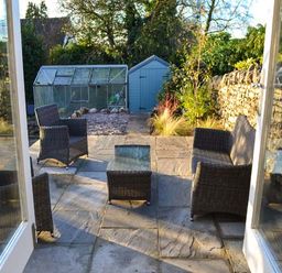 A garden patio with wicker chairs and a glass table in front of a greenhouse and shed at The Potting Shed in Burton-in-Kendal near Kirkby Lonsdale