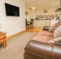 A living area with a sofa and wall mounted TV next to a kitchen and dining table at Snowdrop Cottage in Aldingham