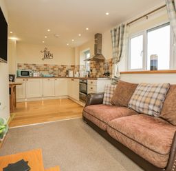 An open plan living room and kitchen with a brown and plaid sofa a wall mounted tv and kitchen cabinets with tiled backsplash at Snowdrop Cottage in Aldingham