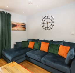 A living room with a large blue sectional sofa with green and orange cushions a wall clock and a window with green curtains at Bluebell Cottage in Aldingham